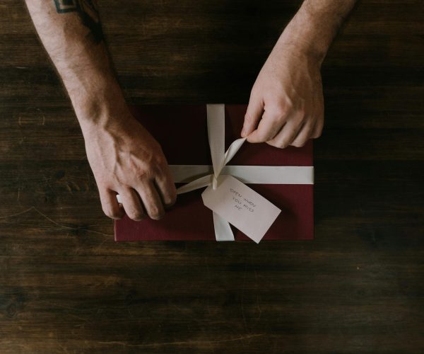 Hands wrapping a red gift box with a white ribbon on a wooden surface, ideal for holiday or celebration themes.